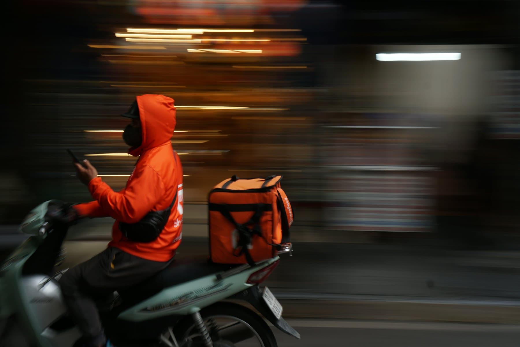 A delivery rider in an orange jacket riding through the city with a delivery box